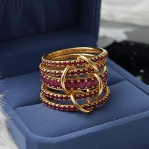K&ouml;nnte beinhalten: Ein mehrschichtiger Goldring mit zahlreichen kleinen, oval geformten roten Edelsteinen. Der Ring befindet sich in einer blauen Schmuckschatulle, die sein kompliziertes Design und seine leuchtende Farbe zeigt. Die Ringb&auml;nder sind durch goldene Schlaufen verbunden.