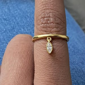 Puede incluir: Un anillo de oro con un colgante de diamante talla marquesa. El anillo es una banda simple con un pequeño lazo que sostiene el diamante. El diamante es transparente y brilla contra el oro.