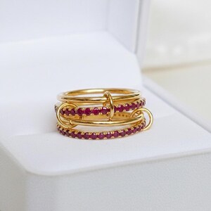 Op de afbeelding: Een goudkleurige ring met drie verbonden banden, waarvan twee bezet zijn met kleine, rode edelstenen. De ring wordt gepresenteerd in een witte doos, die het ontwerp en de kleur benadrukt. Het ontwerp van de ring is modern en elegant.