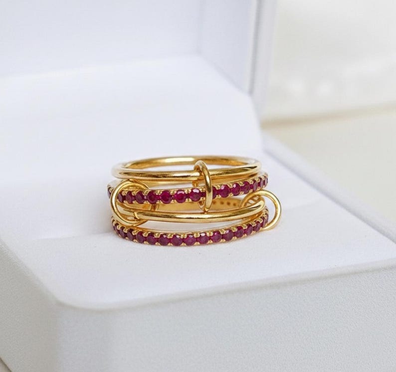 Op de afbeelding: Een goudkleurige ring met drie verbonden banden, waarvan twee bezet zijn met kleine, rode edelstenen. De ring wordt gepresenteerd in een witte doos, die het ontwerp en de kleur benadrukt. Het ontwerp van de ring is modern en elegant.