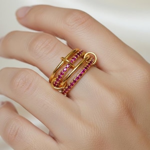 Op de afbeelding: Een goudkleurige ring met drie verbonden banden. Twee banden zijn bezet met kleine, helderrode edelstenen. De ring zit om een vinger. De ring heeft een uniek, modern ontwerp.