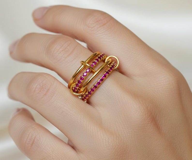 Op de afbeelding: Een goudkleurige ring met drie verbonden banden. Twee banden zijn bezet met kleine, helderrode edelstenen. De ring zit om een vinger. De ring heeft een uniek, modern ontwerp.