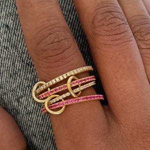 Puede incluir: Un conjunto de cuatro anillos dorados entrelazados con peque&ntilde;os aros dorados. Dos anillos est&aacute;n incrustados con piedras transparentes y dos con piedras rosas. Los anillos est&aacute;n apilados en un dedo sobre un fondo de mezclilla.