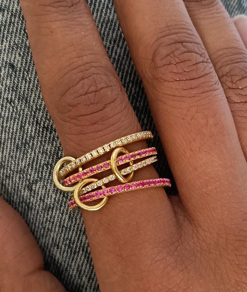 Puede incluir: Un conjunto de cuatro anillos dorados entrelazados con peque&ntilde;os aros dorados. Dos anillos est&aacute;n incrustados con piedras transparentes y dos con piedras rosas. Los anillos est&aacute;n apilados en un dedo sobre un fondo de mezclilla.