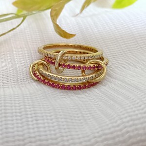 Puede incluir: Un conjunto de anillos dorados con piedras preciosas incrustadas. Algunos anillos tienen piedras transparentes, mientras que otros tienen piedras rojas brillantes. Los anillos est&aacute;n entrelazados, creando un dise&ntilde;o &uacute;nico. Fondo blanco con hojas verdes y amarillas.