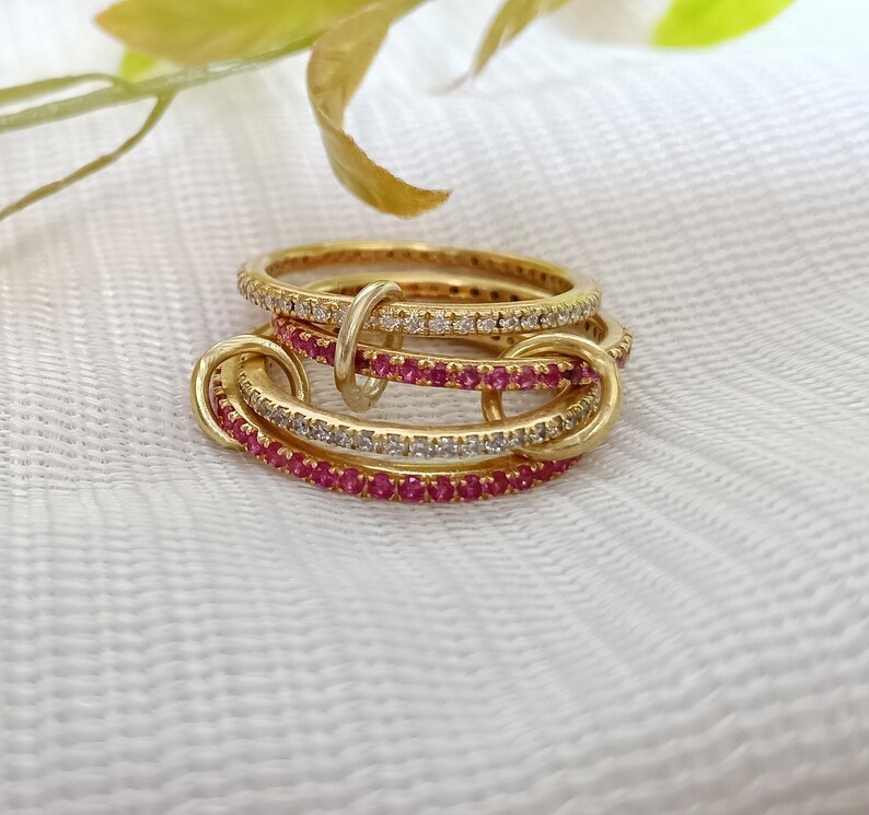Puede incluir: Un conjunto de anillos dorados con piedras preciosas incrustadas. Algunos anillos tienen piedras transparentes, mientras que otros tienen piedras rojas brillantes. Los anillos est&aacute;n entrelazados, creando un dise&ntilde;o &uacute;nico. Fondo blanco con hojas verdes y amarillas.
