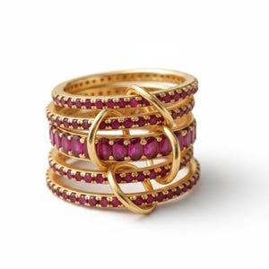 K&ouml;nnte beinhalten: Ein Stapel goldener Ringe mit rubinroten Edelsteinen. Die Ringe sind durch goldene Schlaufen verbunden, wodurch ein einzigartiges Design entsteht. Die Edelsteine sind gleichm&auml;&szlig;ig um jeden Ring angeordnet, und das Gold hat eine polierte Oberfl&auml;che.