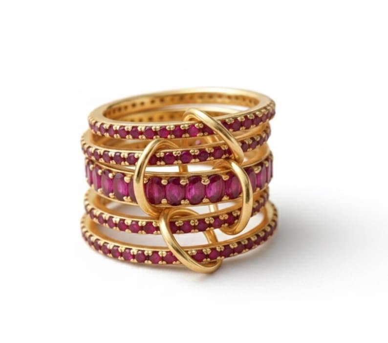 K&ouml;nnte beinhalten: Ein Stapel goldener Ringe mit rubinroten Edelsteinen. Die Ringe sind durch goldene Schlaufen verbunden, wodurch ein einzigartiges Design entsteht. Die Edelsteine sind gleichm&auml;&szlig;ig um jeden Ring angeordnet, und das Gold hat eine polierte Oberfl&auml;che.