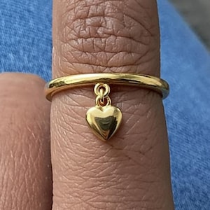 May include: A gold-toned ring featuring a smooth, polished band with a small, dangling heart-shaped charm attached to the centre. The heart charm is solid and reflective, hanging from a tiny circular link connected directly to the ring band.