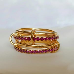 Op de afbeelding: Een stapelbare set gouden ringen, verbonden door gouden lussen en een centrale ring. Elke ring is versierd met kleine, rode edelstenen, wat een luxueus ontwerp cre&euml;ert. De ringen staan tegen een zachte, neutrale achtergrond.