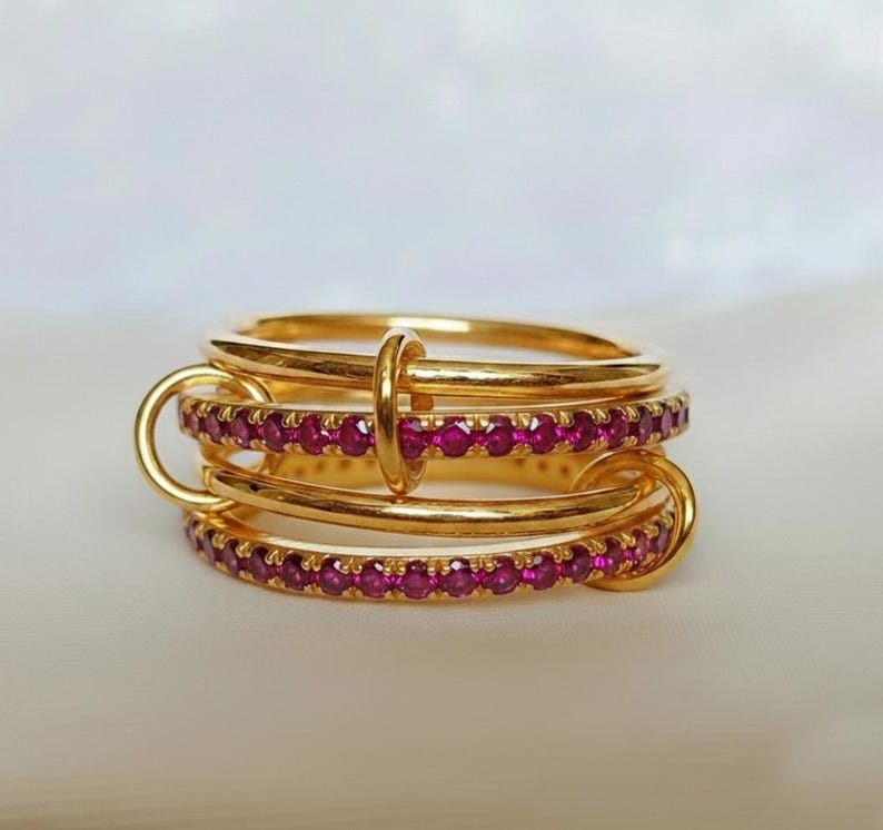 Op de afbeelding: Een stapelbare set gouden ringen, verbonden door gouden lussen en een centrale ring. Elke ring is versierd met kleine, rode edelstenen, wat een luxueus ontwerp cre&euml;ert. De ringen staan tegen een zachte, neutrale achtergrond.