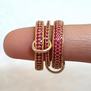 May include: Three gold-coloured rings, each set with small, bright pink gemstones. The rings are stacked together, with a smaller ring in the middle. The rings are held by a finger against a plain white background.