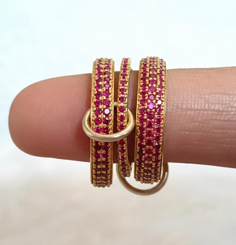 May include: Three gold-coloured rings, each set with small, bright pink gemstones. The rings are stacked together, with a smaller ring in the middle. The rings are held by a finger against a plain white background.