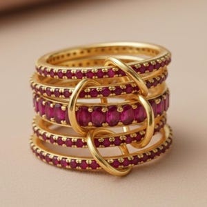 K&ouml;nnte beinhalten: Ein Stapel goldfarbener Ringe mit einem Band aus leuchtend rubinroten Edelsteinen. Die Ringe sind mit goldenen B&ouml;gen verbunden, wodurch ein einzigartiges Design entsteht. Der Schmuck wird auf einem neutralen Hintergrund pr&auml;sentiert.