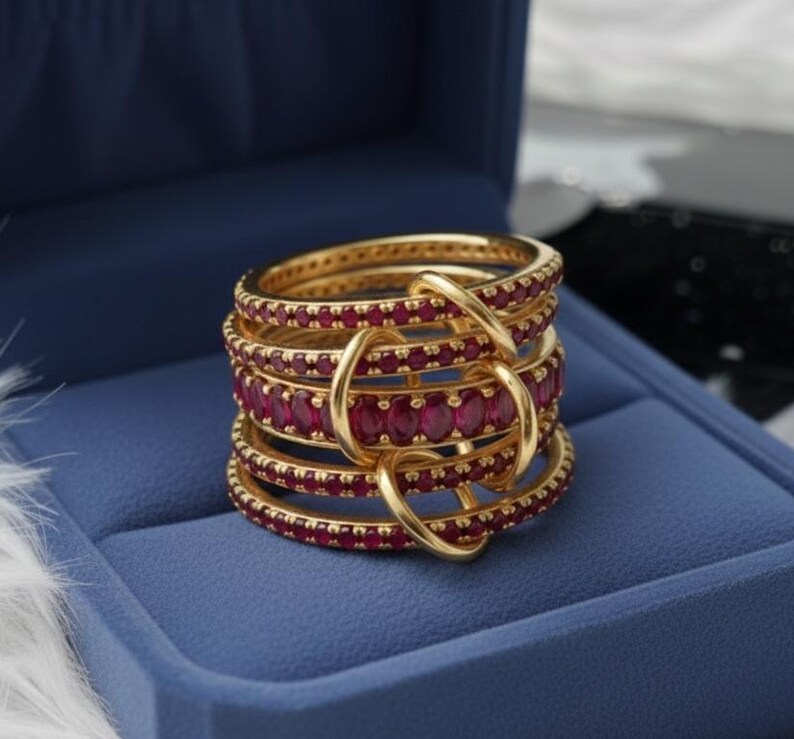 K&ouml;nnte beinhalten: Ein goldener Ring mit mehreren Reihen, besetzt mit kleinen, ovalen roten Edelsteinen. Der Ring wird in einer blauen Schatulle pr&auml;sentiert. Das Design weist ineinandergreifende Elemente auf und ist ein auff&auml;lliges Schmuckst&uuml;ck.