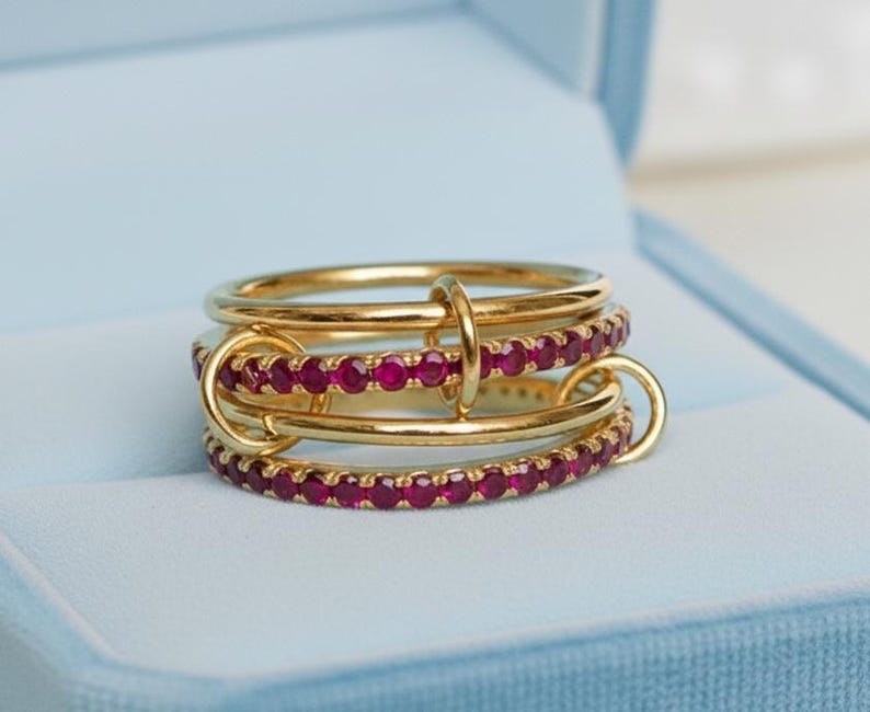 Op de afbeelding: Een goudkleurige ring met drie banden, waarvan &eacute;&eacute;n met een rij kleine, rode edelstenen. De banden zijn verbonden met kleine gouden ringen. De ring wordt gepresenteerd in een lichtblauwe sieradendoos.