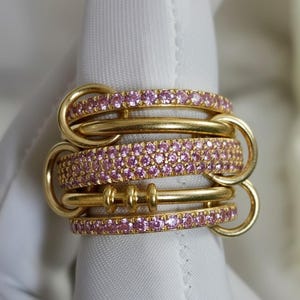 Puede incluir: Un anillo de oro de varias capas con piedras preciosas rosas. El anillo presenta varias bandas conectadas por bucles de oro. Las bandas están adornadas con pequeñas piedras preciosas rosas muy juntas, creando un efecto brillante. El oro tiene un acabado pulido.