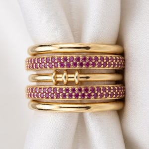Può includere: Una pila di cinque anelli d'oro. Gli anelli superiore e inferiore sono fasce in oro lucido liscio. Due anelli presentano file di piccole pietre rotonde di colore rosa vibrante. L'anello centrale è una fascia d'oro liscia con tre piccole barre verticali.