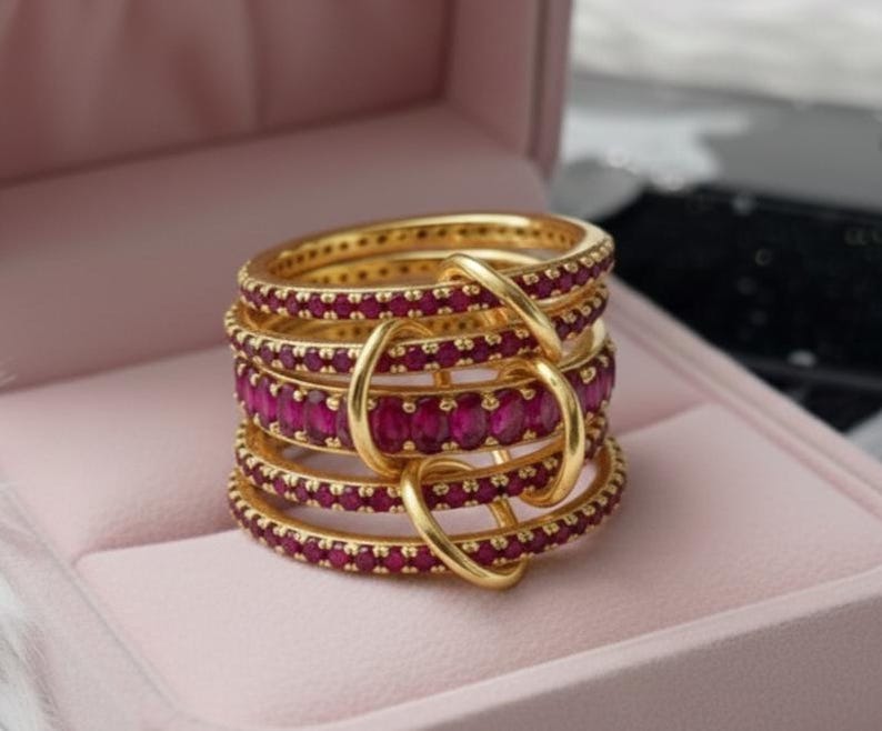 K&ouml;nnte beinhalten: Ein Stapel goldfarbener Ringe mit einem Band aus leuchtend rubinroten Edelsteinen. Die Ringe sind mit goldenen Schlaufen verbunden, wodurch ein einzigartiges Design entsteht. Die Ringe werden in einer rosa Schmuckschatulle pr&auml;sentiert.
