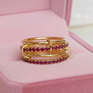 Op de afbeelding: Een goudkleurige ring met drie banden, elk versierd met kleine, rode edelstenen. De banden zijn verbonden door gouden lussen. De ring wordt gepresenteerd in een roze sieradendoos.