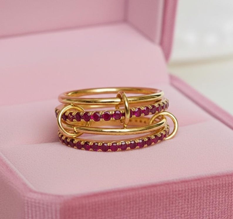 Op de afbeelding: Een goudkleurige ring met drie banden, elk versierd met kleine, rode edelstenen. De banden zijn verbonden door gouden lussen. De ring wordt gepresenteerd in een roze sieradendoos.