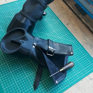 Puede incluir: Dos protectores de brazo de cuero negro con hebillas y correas. Uno está en posición vertical, el otro está abierto, revelando el interior. Una pequeña herramienta con mango de madera descansa sobre el protector abierto. Los protectores de brazo están sobre una esterilla de corte verde.