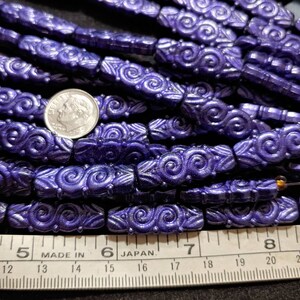 May include: A close-up of a pile of purple glass beads with a swirl design. The beads are approximately 1.2 cm long and are arranged in rows. A ruler is visible in the bottom of the image, with the numbers 5, 6, 7, and 8 visible. The ruler is marked "Made in" and "Japan."
