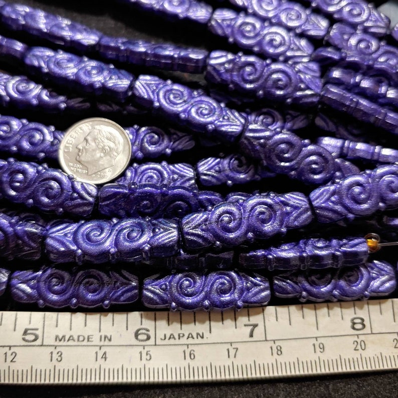 May include: A close-up of a pile of purple glass beads with a swirl design. The beads are approximately 1.2 cm long and are arranged in rows. A ruler is visible in the bottom of the image, with the numbers 5, 6, 7, and 8 visible. The ruler is marked "Made in" and "Japan."