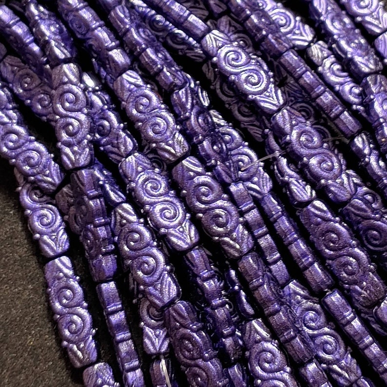 May include: A close-up of a strand of purple glass beads with a swirl design. The beads are all the same size and shape, and they are arranged in a row.