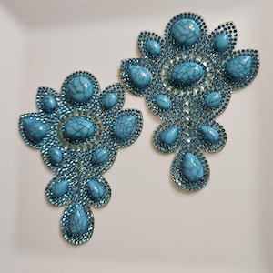 May include: Two turquoise-colored, ornate appliques. Each applique features a central oval stone surrounded by smaller stones and teardrop-shaped accents. The edges are lined with small, sparkling rhinestones, creating a decorative floral design.