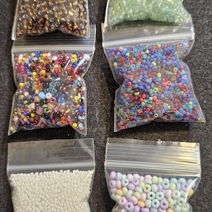 May include: Six clear plastic bags filled with various seed beads. Colors include gold, green, mixed colors, blue, white, and pastel. The beads are small and round, suitable for jewelry making and crafts.