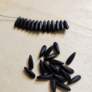 May include: A close-up of black glass beads in the shape of teardrops. The beads are scattered on a wooden surface.