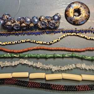 May include: A collection of different types of beads, including blue glass beads, brown wooden beads, white stone beads, green glass beads, and a black and gold patterned bead. The beads are arranged in rows on a gray surface.