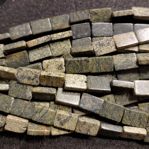 May include: A close-up of a pile of green and brown rectangular gemstone beads. The beads have a mottled, speckled appearance.