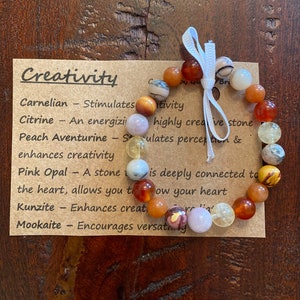 May include: A brown bracelet with various colored stones, including carnelian, citrine, peach aventurine, pink opal, kunzite, and mookaite. The bracelet is tied with a white ribbon and is accompanied by a card that reads "Creativity" and lists the properties of each stone.