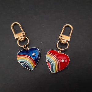 May include: Two gold keyrings with heart-shaped charms. One charm is blue with a rainbow design and the other is red with a rainbow design. Both charms have a small rhinestone in the centre.