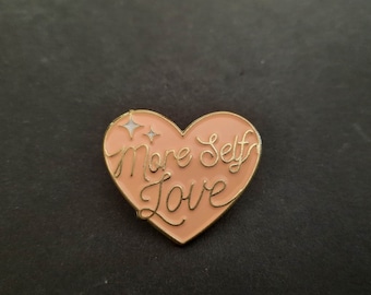 Könnte beinhalten: Ein herzförmiger Emaille-Anstecker in einem sanften Pfirsichton mit goldenem Rand. Der Anstecker trägt die Worte "More Self Love" in goldener Schrift sowie zwei kleine Sternenmotive. Auf dunklem Hintergrund.