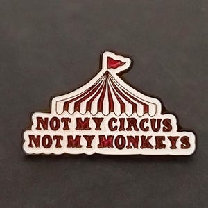 Könnte beinhalten: Eine Metall-Emaille-Anstecknadel in Form eines Zirkuszeltes mit rot-weißen Streifen und einer kleinen roten Flagge an der Spitze. Der Text NOT MY CIRCUS NOT MY MONKEYS ist in dunkelroten Großbuchstaben auf weißem Grund am Zeltfuß gedruckt.