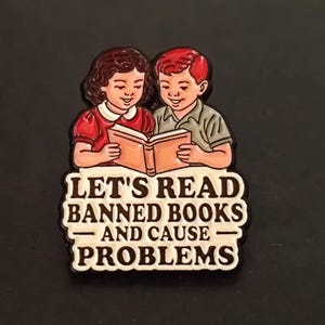 Peut inclure: Un badge représentant deux enfants lisant un livre. Les enfants sont dessinés dans un style rétro, l'un portant une robe rouge et l'autre une chemise verte. Le texte sur le badge se lit "LET'S READ BANNED BOOKS AND CAUSE PROBLEMS."