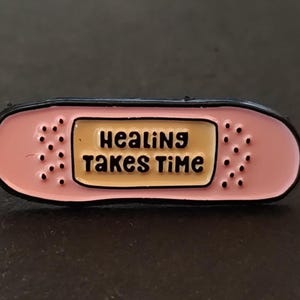 Peut inclure: Épingle en émail rose en forme de pansement avec l'inscription noire « Healing Takes Time ». L'épingle a un bord noir et de petits détails en pointillés.