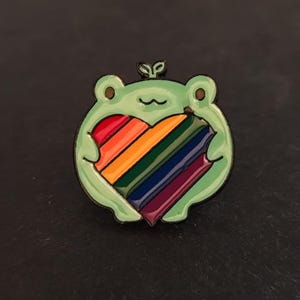 Puede incluir: Un pin de esmalte verde con forma de rana que sostiene un corazón arcoíris. El pin tiene un contorno negro y un pequeño brote en la parte superior de la cabeza de la rana. El corazón presenta rayas rojas, naranjas, amarillas, verdes, azules y moradas.