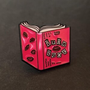 Peut inclure: Une épingle en émail rose en forme de livre ouvert. L'épingle présente le texte "Burn Book" avec des graphiques de lèvres et de baisers. Les pages du livre sont blanches et l'épingle a un contour noir.