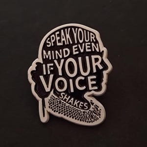 Op de afbeelding: Een zwart-witte emaille pin in de vorm van een hoofd met baard. De pin bevat de tekst "SPEAK YOUR MIND EVEN IF YOUR VOICE SHAKES" in een vet, leesbaar lettertype. De pin heeft een zilveren rand en staat tegen een donkere achtergrond.