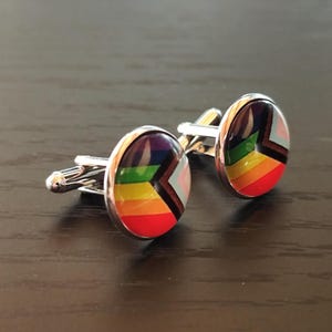 Peut inclure: Une paire de boutons de manchette argentés avec un motif de drapeau arc-en-ciel coloré. Les boutons de manchette ronds ont une finition brillante, avec les couleurs de l'arc-en-ciel disposées en chevrons. Le métal est brillant.