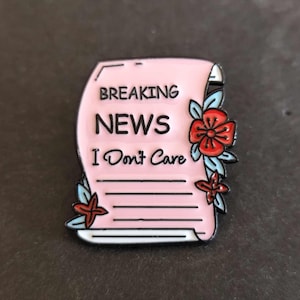 Puede incluir: Pin de esmalte rosa con un diseño de rollo que dice "Breaking News I Don't Care" con flores rojas y azules.