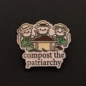 Può includere: Una spilla in smalto metallico raffigurante tre gatti bianchi con cappelli di paglia e salopette verdi, attorno a una compostiera in legno. Il gatto centrale tiene un forcone. Il testo "compost the patriarchy" è stampato in marrone scuro in basso.