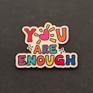 Op de afbeelding: Een metalen emaille speld met de tekst YOU ARE ENOUGH in kleurrijke, ronde letters. Het woord YOU is in rood en oranje met een roze hartje dat de letter O vervangt. De woorden ARE ENOUGH zijn in paars, groenblauw, oranje, geel, rood en blauw.