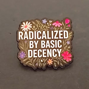Peut inclure: Une broche en émail noir à bord festonné portant le texte blanc RADICALIZED BY BASIC DECENCY au centre. Le motif est entouré d'une illustration botanique colorée composée de diverses petites fleurs et feuilles roses, blanches et violettes.