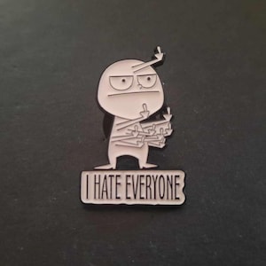May include: A white enamel pin depicting an irate cartoon figure, displaying multiple middle fingers. The pin includes the text "I HATE EVERYONE" on a rectangular banner. The pin is set against a dark background.