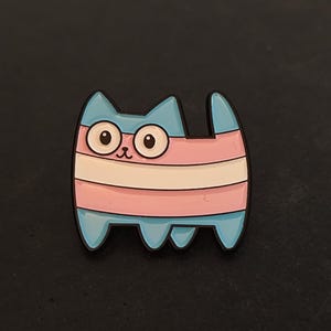 Puede incluir: Un pin de gato de dibujos animados con un contorno azul y un diseño con los colores de la bandera transgénero: azul claro, rosa y blanco. El gato tiene ojos grandes y redondos y una nariz pequeña. El pin es de metal.